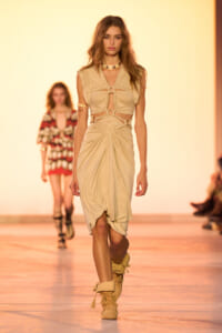 Model walking the runway in a beige cut-out dress with knotted bodice and draped skirt, paired with beige boots and chunky jewelry.