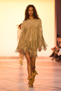 Model walks the runway in a beige fringe dress and matching boots on an orange-lit catwalk.