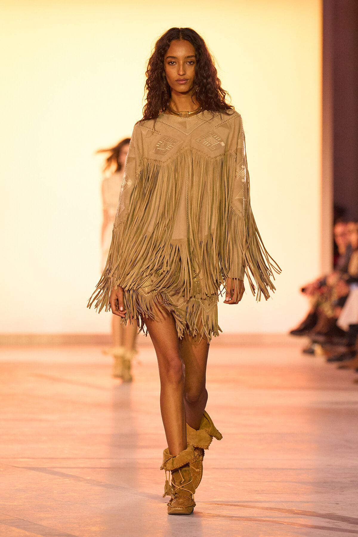 Model walks the runway in a beige fringe dress and matching boots on an orange-lit catwalk.
