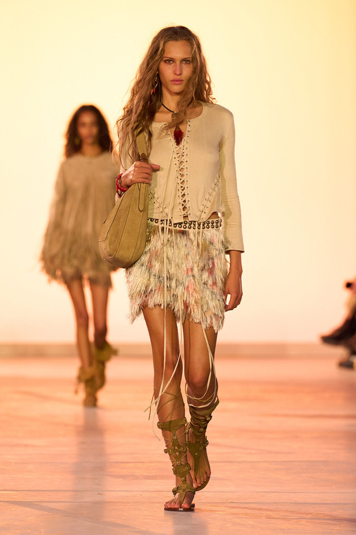 Model walking a fashion runway in a beige lace-up top and a pastel feathered mini skirt, carrying a tan handbag and wearing tan gladiator sandals.