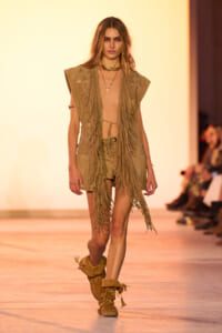 Model on a runway wearing a tan suede fringe vest over a bare torso, matching shorts, and tan fringe boots; warm backdrop behind.