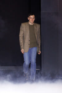 Man walks out from fog on a dark stage, wearing a brown plaid blazer, olive green shirt, and blue jeans.