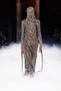 Model on a runway wearing a silver beaded fringe gown, surrounded by stage fog.