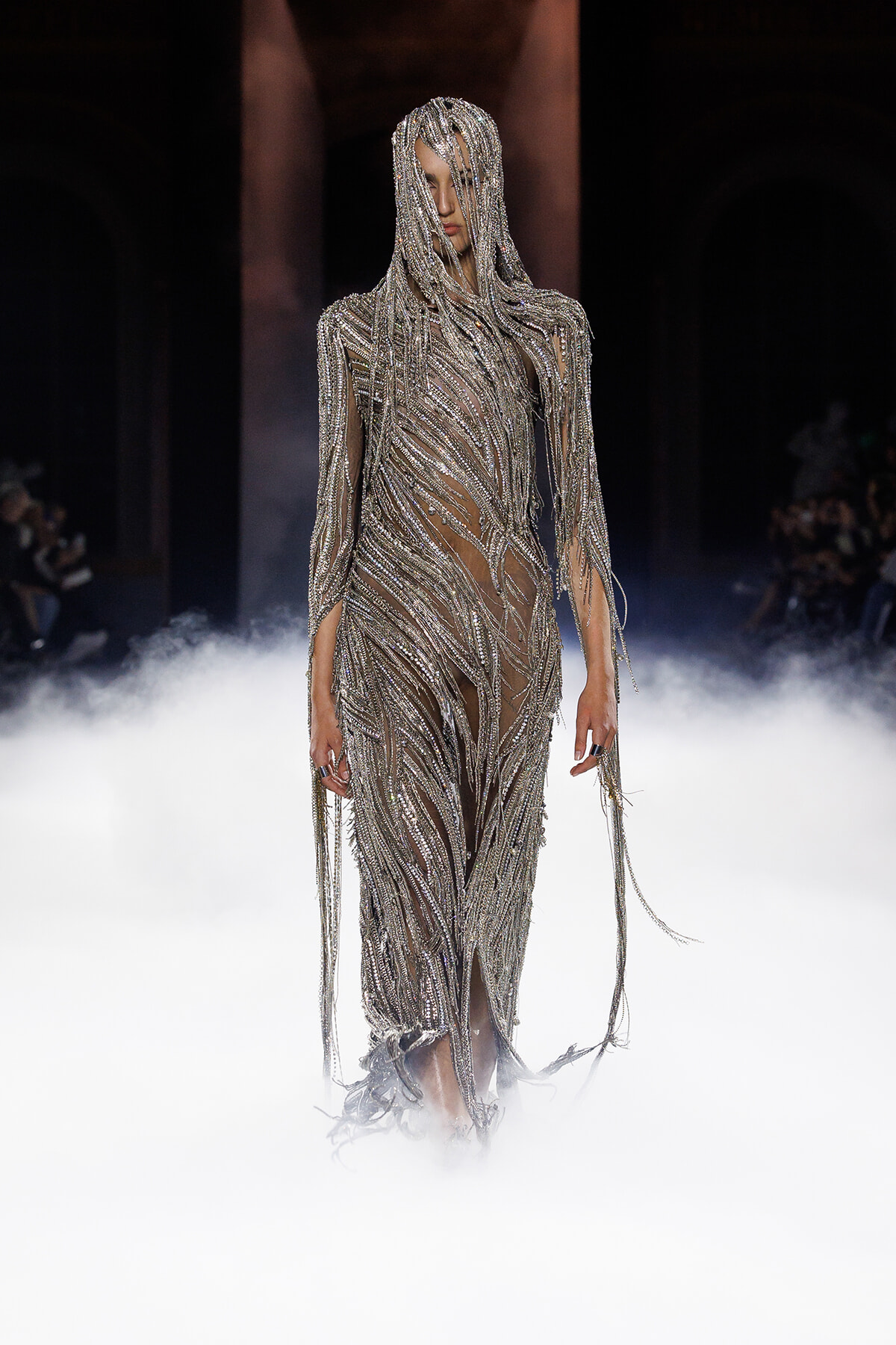 Model on a runway wearing a silver beaded fringe gown, surrounded by stage fog.