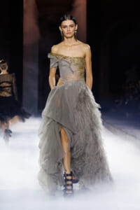 Model in a light gray off-shoulder gown with gold embroidery and a feathered, layered train on a runway.