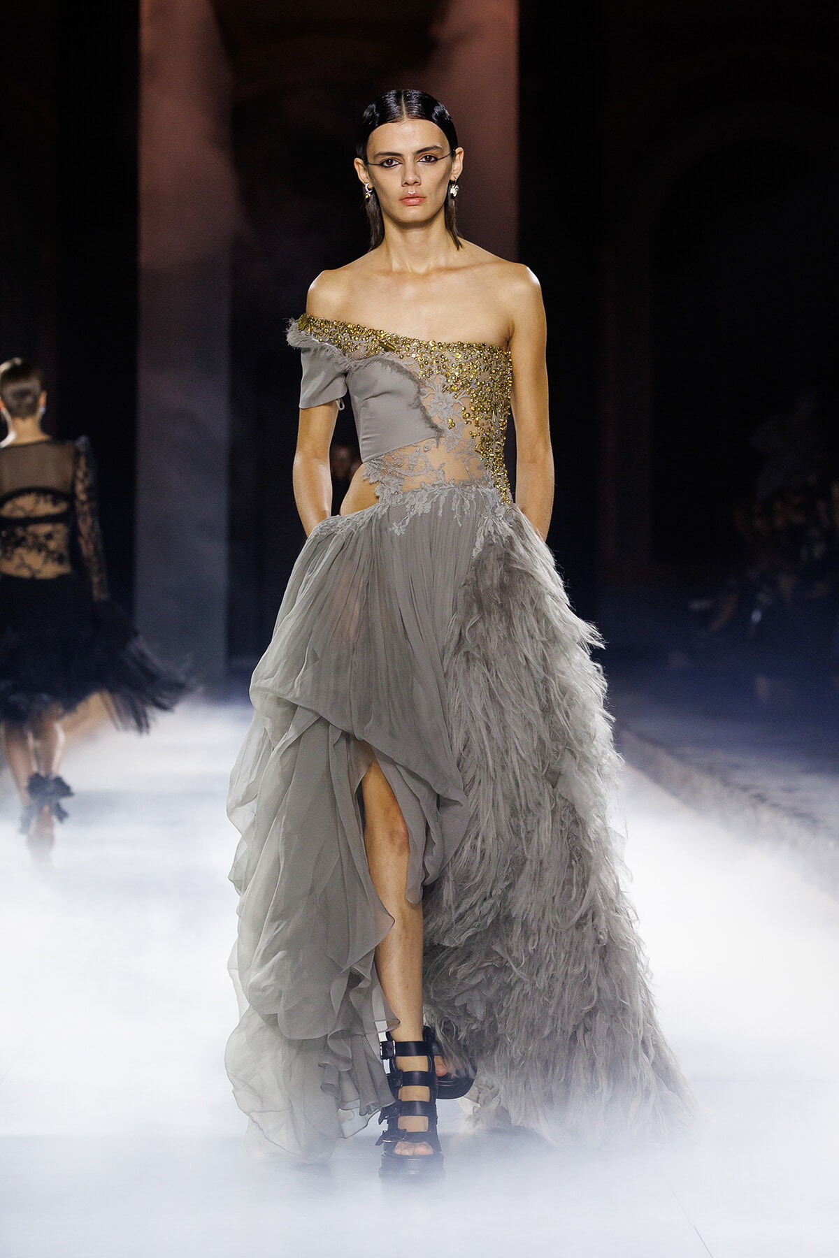 Model in a light gray off-shoulder gown with gold embroidery and a feathered, layered train on a runway.