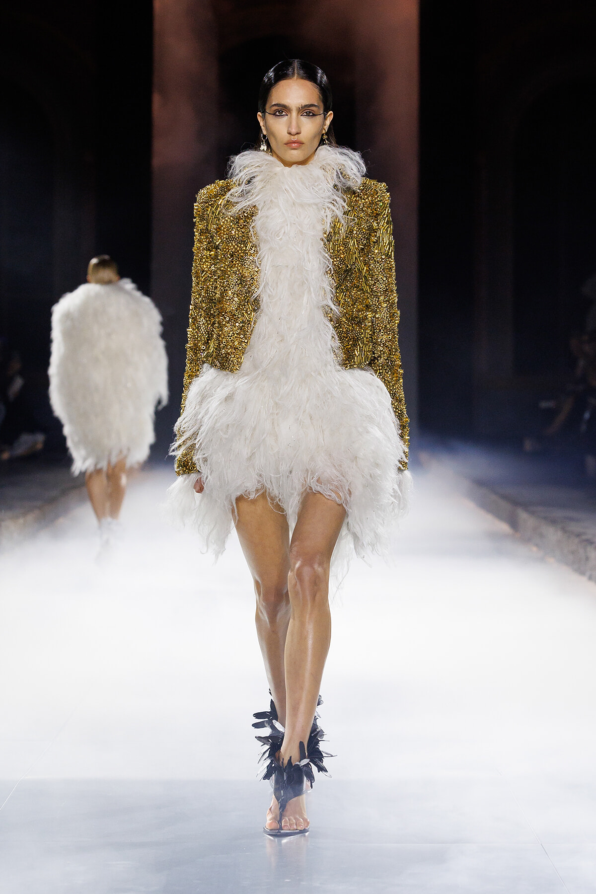 Model walks the runway wearing a gold sequin jacket over a white feathered dress; foggy catwalk.