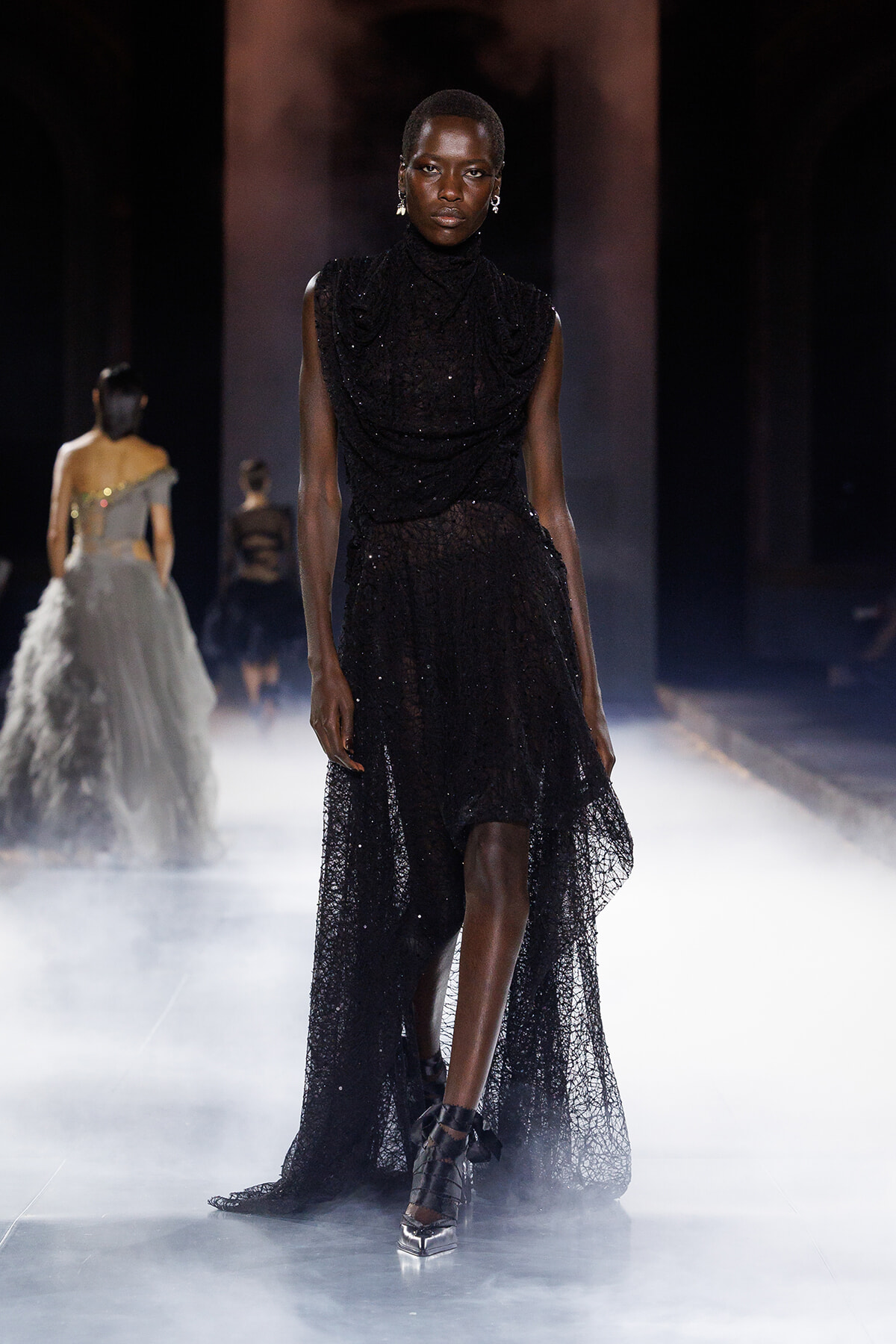 Runway model in a black sequined sleeveless gown with a high-low lace skirt walking through fog.