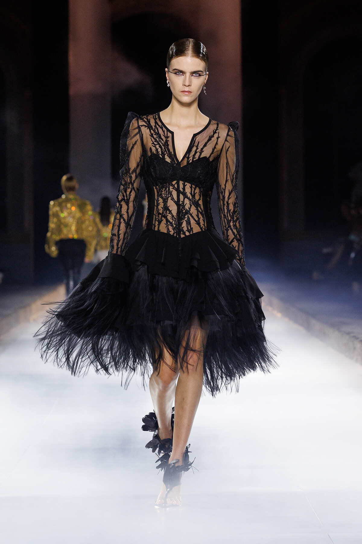 Model walks the runway in a black sheer lace gown with a structured bodice and layered feathered skirt.