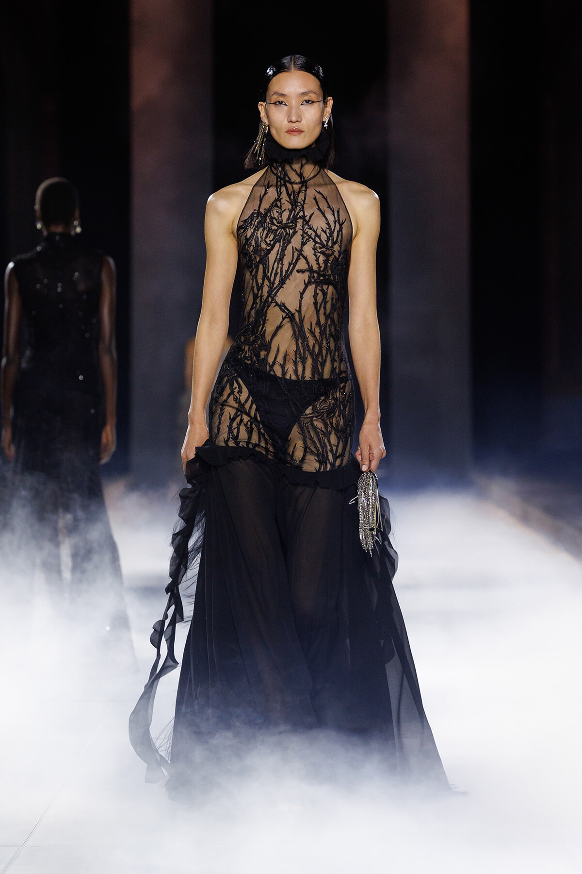 Model walking a runway in a sheer black gown with branch-like embroidery and a flowing skirt, holding a silver tassel clutch.