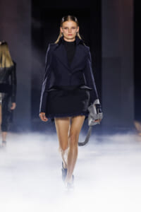 Model on a foggy runway wearing a navy tailored blazer, black top and mini skirt, holding a gray handbag with fringe tassels.