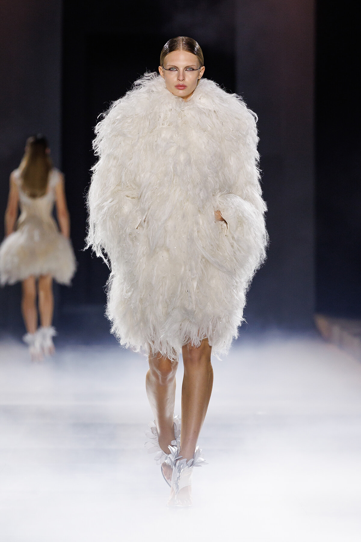 Model walking a runway in a voluminous white feathered coat, with a sleek hairstyle and dramatic makeup.