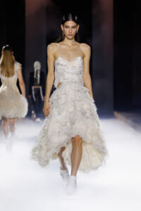 Model walks a white strapless, ruffled gown on a foggy runway with dramatic lighting.