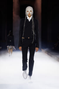 Model walks a foggy runway in a black tuxedo with a beaded white mask and dangling chains, high-fashion look for a show.