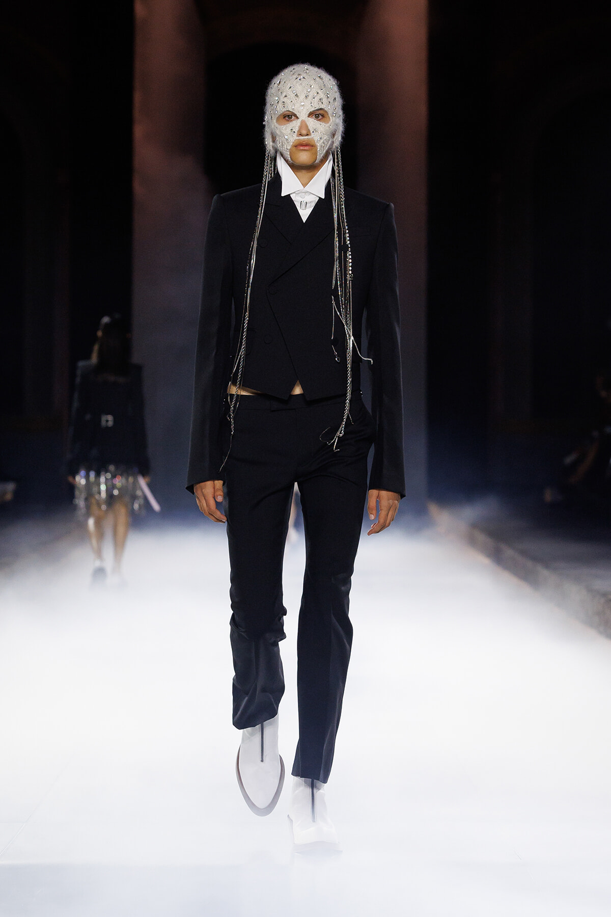 Model walks a foggy runway in a black tuxedo with a beaded white mask and dangling chains, high-fashion look for a show.