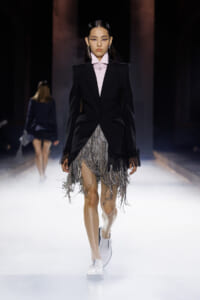 Runway model in a black blazer over a pale pink shirt with a silver fringe skirt and white loafers, walking a spotlighted catwalk.