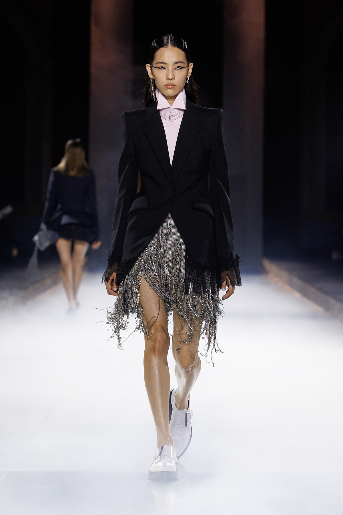 Runway model in a black blazer over a pale pink shirt with a silver fringe skirt and white loafers, walking a spotlighted catwalk.