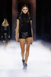 Model walking a runway in a sheer black top, sleeveless vest, and a short black skirt with large buttons, under stage lighting.
