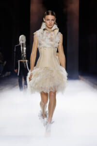Model walks the runway in a cream, feathered mini dress with lace bodice and high collar.