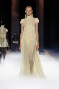 Model walks the runway in a sheer pale yellow high-neck gown with short sleeves and a flowing skirt.