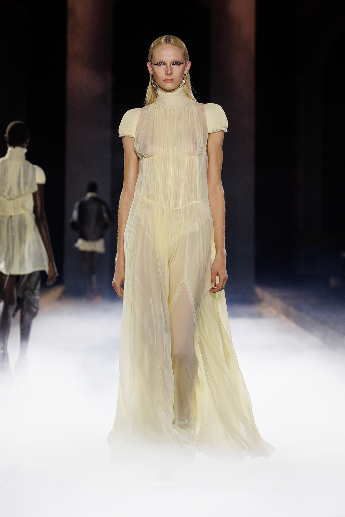 Model walks the runway in a sheer pale yellow high-neck gown with short sleeves and a flowing skirt.