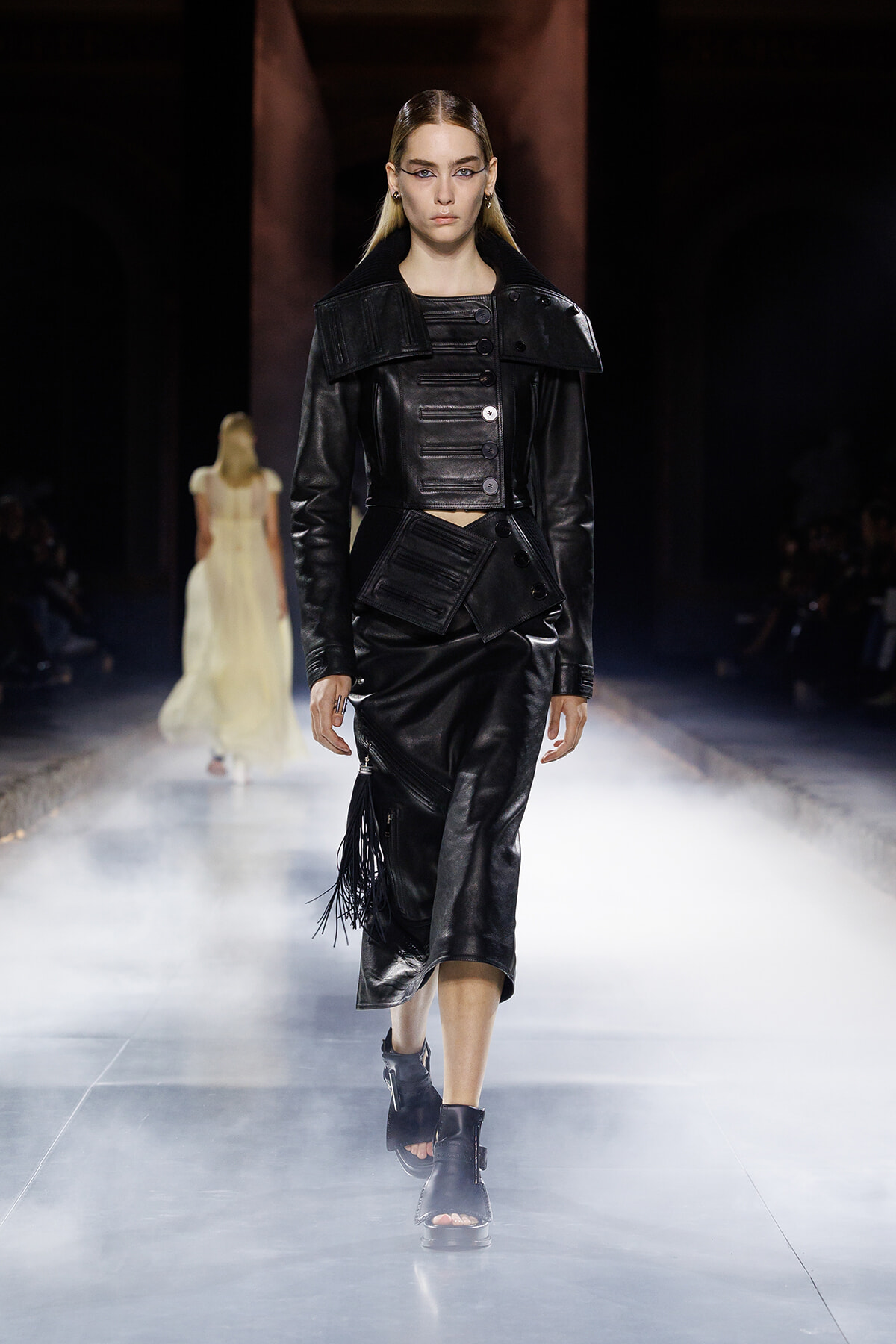 Model on a runway wearing a black, structured leather cropped jacket with a fringe-accented skirt and tall collar details.