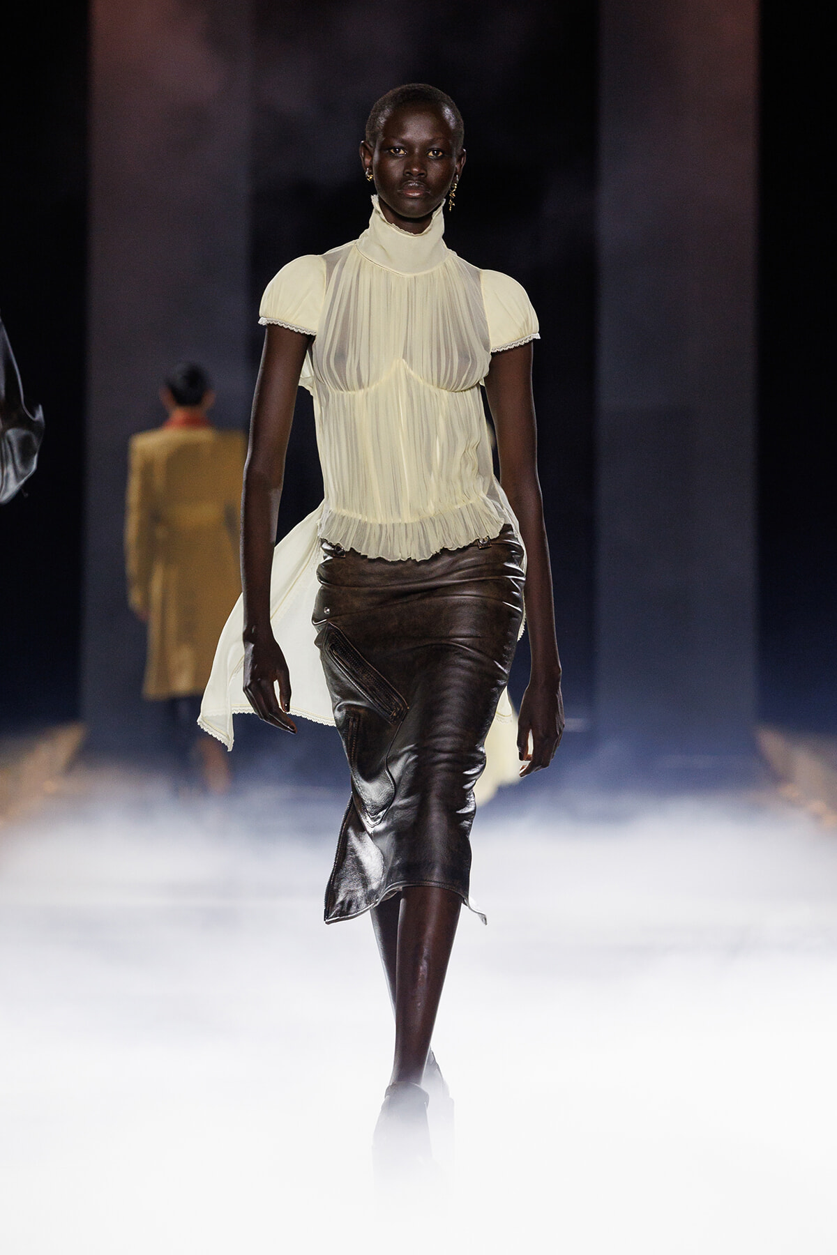 Model walking a fashion runway in a sheer ivory high-neck blouse and dark leather midi skirt with foggy catwalk lights behind.