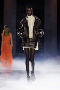 Model walks the smoky runway in a dark brown leather biker jacket over a cream pleated high-neck dress, black tights, and ankle boots.
