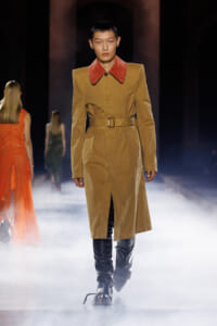 Model walking the runway in a tan belted trench with a red oversized collar and black knee‑high boots amid fog.