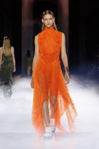 Model on a runway wearing a bright orange, draped lace high-low dress with a turtleneck, white sandals, and black cuffs with futuristic styling.