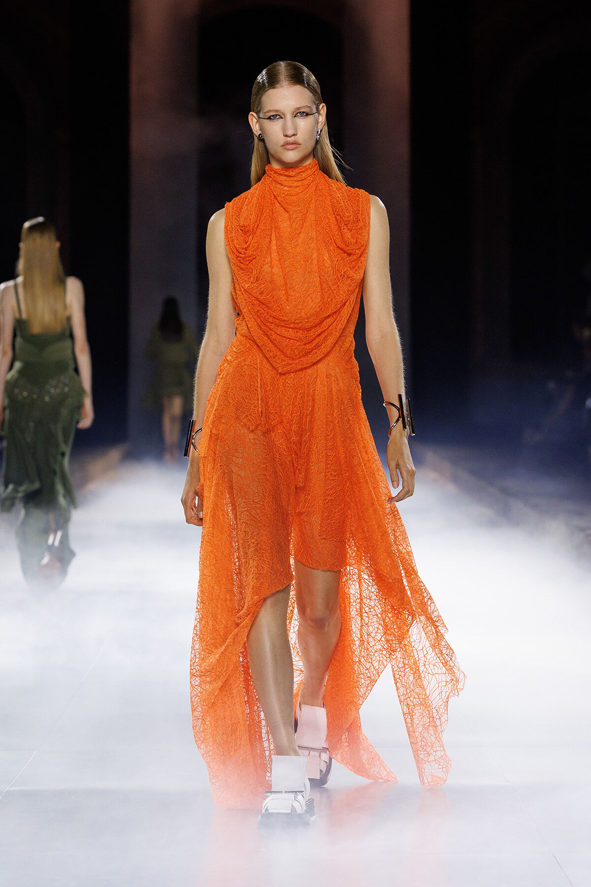 Model on a runway wearing a bright orange, draped lace high-low dress with a turtleneck, white sandals, and black cuffs with futuristic styling.