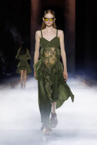 Model walking a foggy runway in an emerald green, sheer-lace gown with floral appliqué and spaghetti straps, wearing yellow sunglasses.