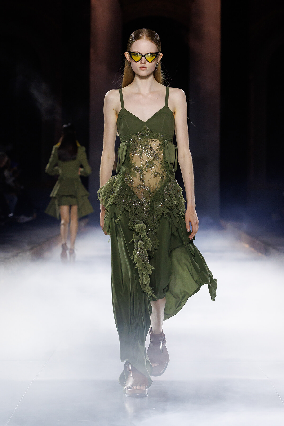 Model walking a foggy runway in an emerald green, sheer-lace gown with floral appliqué and spaghetti straps, wearing yellow sunglasses.