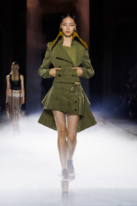 Model walking the runway in an olive-green double-breasted jacket and asymmetrical mini skirt, on a foggy catwalk.