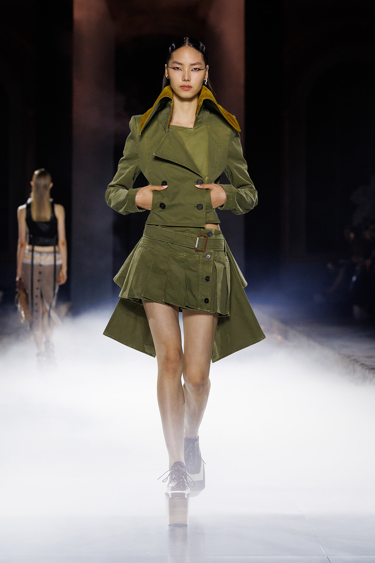 Model walking the runway in an olive-green double-breasted jacket and asymmetrical mini skirt, on a foggy catwalk.