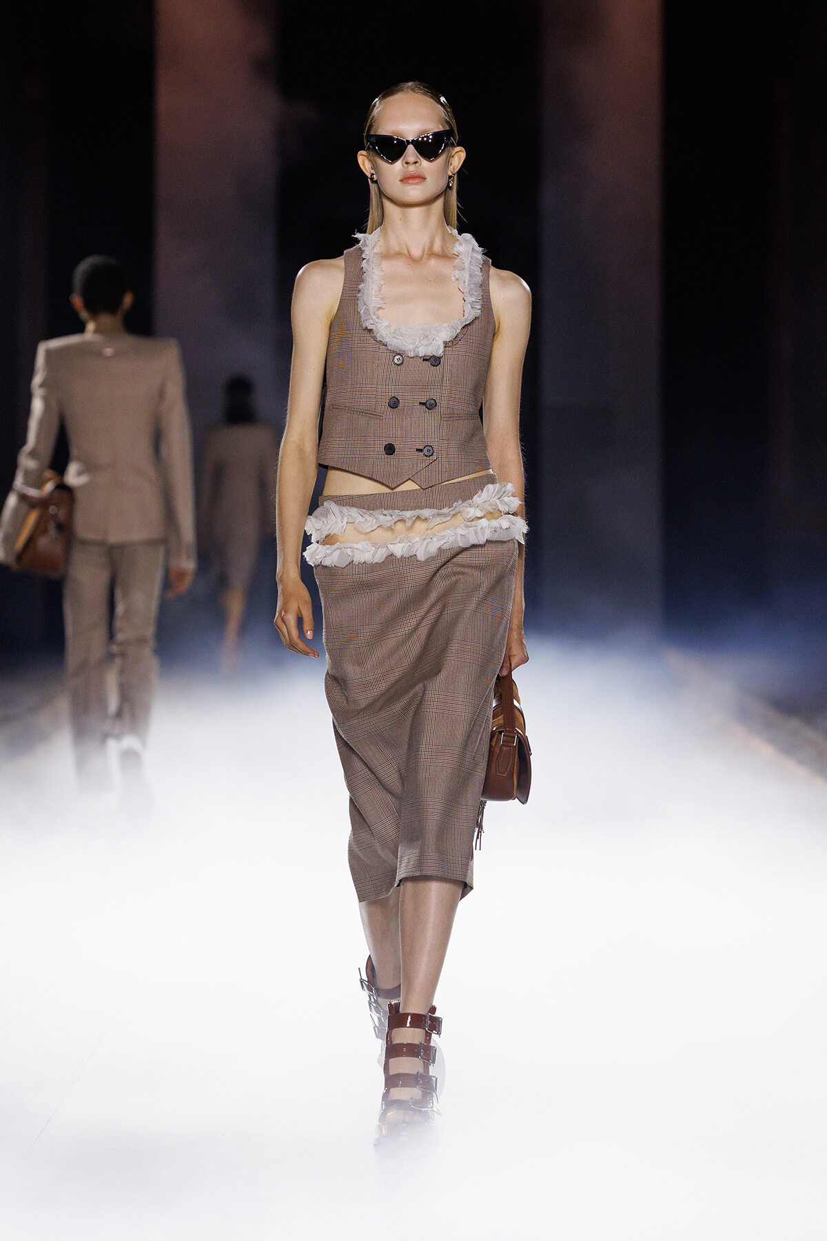 Model walks a foggy runway in a brown plaid sleeveless vest and matching skirt with white ruffle trim, carrying a brown handbag and wearing sunglasses.
