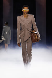 Runway model wearing a tan plaid suit with oversized blazer and matching pants, holding a brown leather bag, and yellow angular sunglasses amid foggy stage.