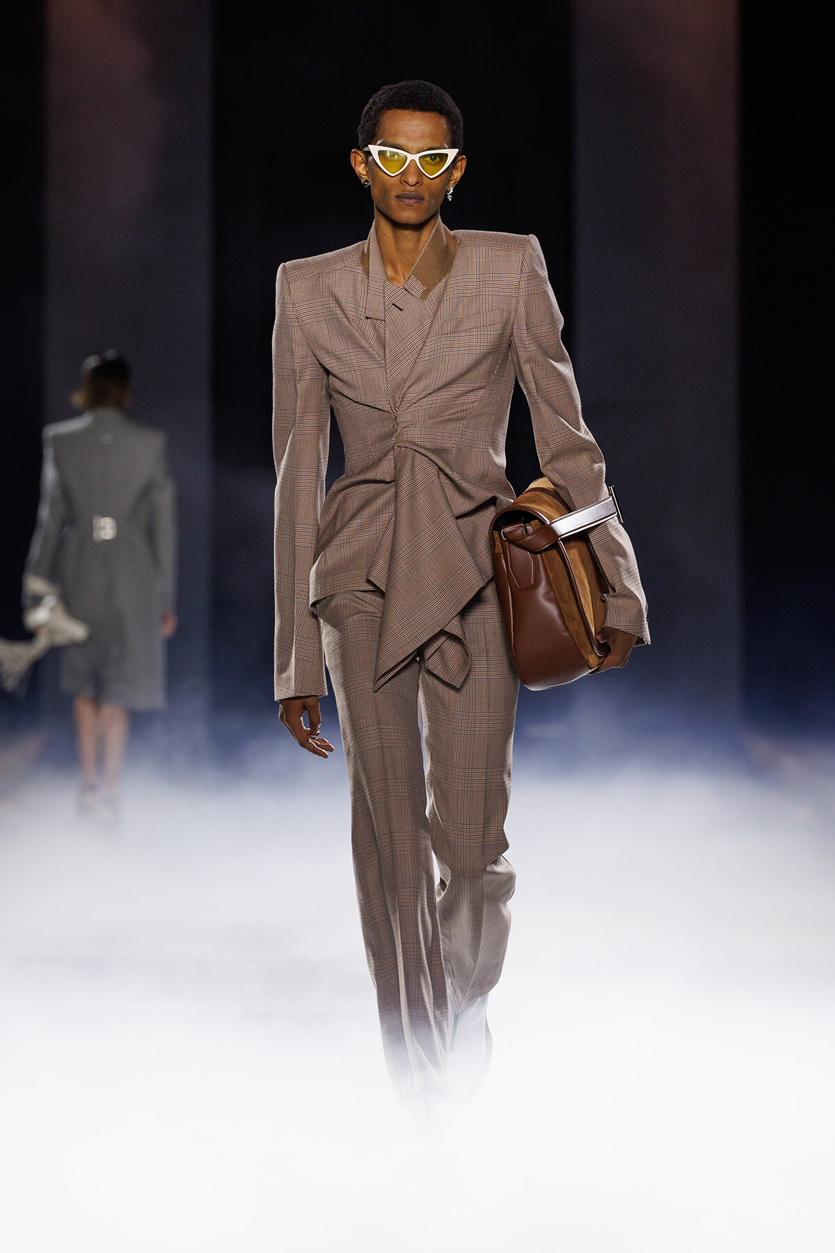 Runway model wearing a tan plaid suit with oversized blazer and matching pants, holding a brown leather bag, and yellow angular sunglasses amid foggy stage.