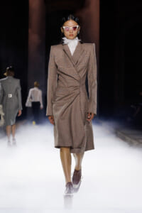 Model on a runway wearing a brown plaid, wrap-style blazer dress with structured shoulders and a white ruffled collar, plus white cat-eye sunglasses.