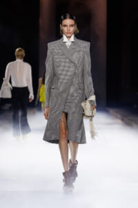 Model walks a foggy runway in a gray checkered double-breasted coat, carrying a white fringe bag.