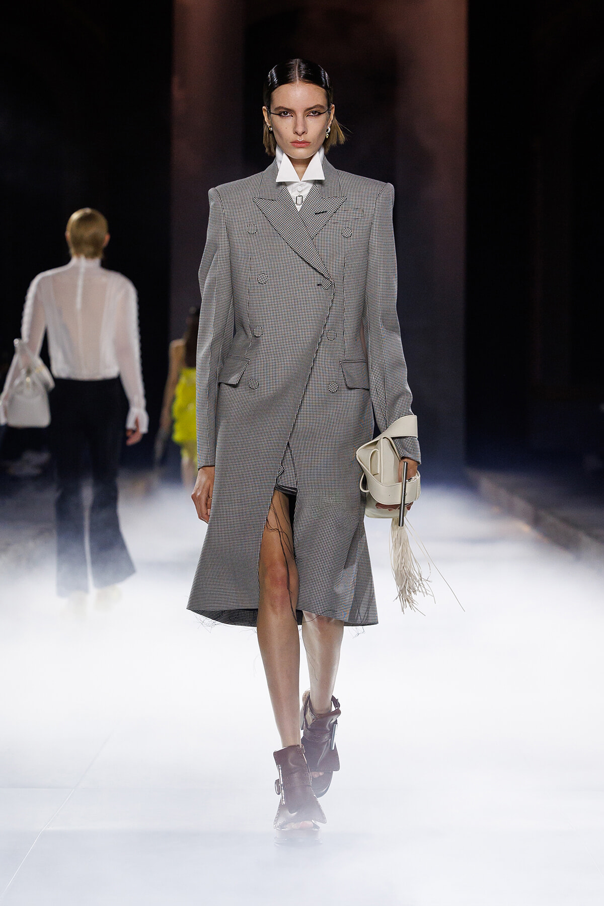 Model walks a foggy runway in a gray checkered double-breasted coat, carrying a white fringe bag.