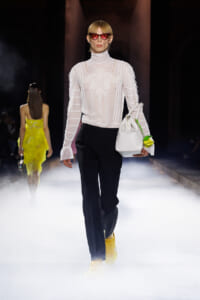 Model walks the runway in a sheer white lace blouse, black wide-leg pants, yellow sneakers, and red cat-eye sunglasses, carrying a white bag with a neon green flower charm