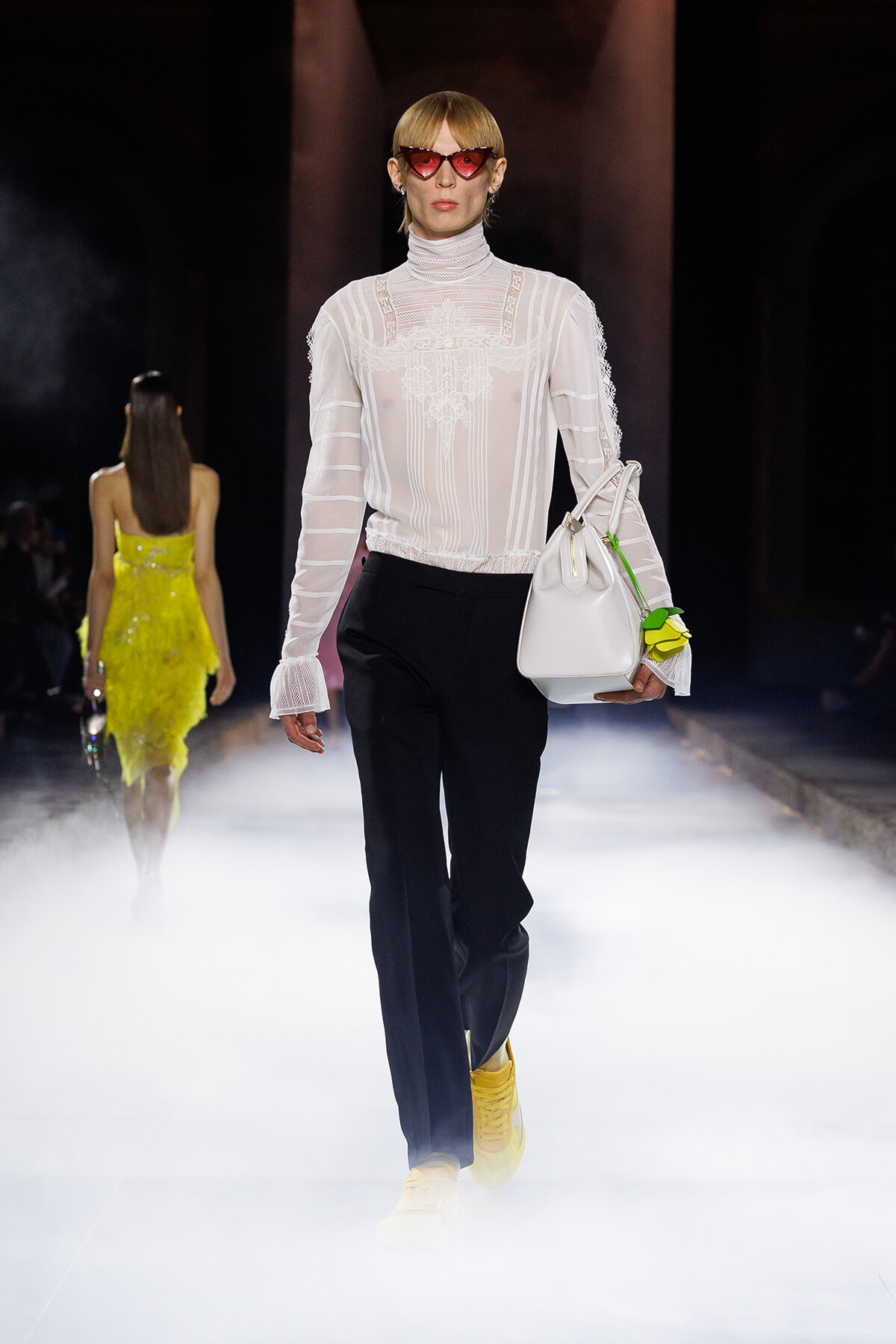 Model walks the runway in a sheer white lace blouse, black wide-leg pants, yellow sneakers, and red cat-eye sunglasses, carrying a white bag with a neon green flower charm