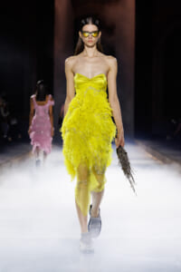 Model walks a runway in a bright yellow strapless dress with feathered skirt and metallic sunglasses, holding a fringe clutch.