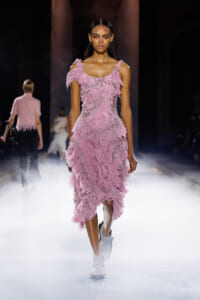 Model walking a runway in a pink fringe evening gown with silver beading and a fitted bodice, surrounded by fog on the catwalk