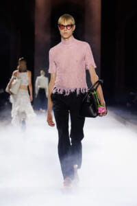 Model walking the runway in a pink lace top, black pants, pink sunglasses, and a black handbag with a pink flower accessory.