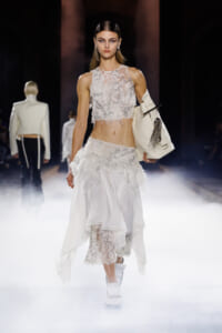 Model in a white lace crop top and layered white skirt with fringe, carrying a large white tote on a foggy runway.