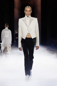 Model walks a fashion show wearing a white cropped double-breasted blazer, white shirt, and black trousers amid stage fog.