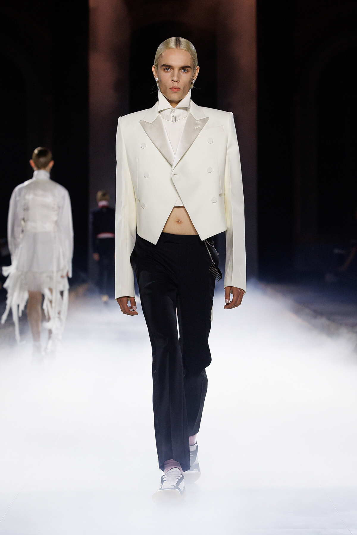 Model walks a fashion show wearing a white cropped double-breasted blazer, white shirt, and black trousers amid stage fog.