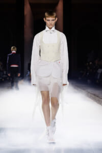 Model walks a fashion runway in an off-white structured vest over a white blouse with long sleeves and a sheer, tiered skirt, surrounded by mist.